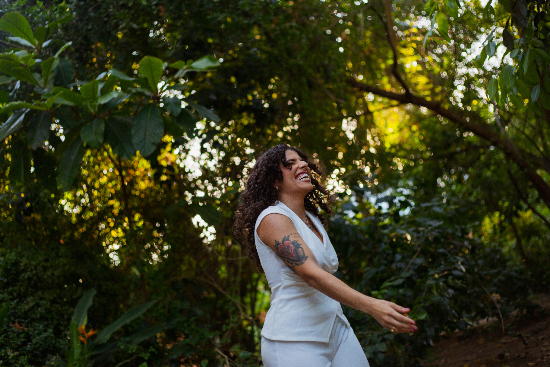 Laisla sorrindo em meio à vegetação, com luz natural filtrada pelas folhas